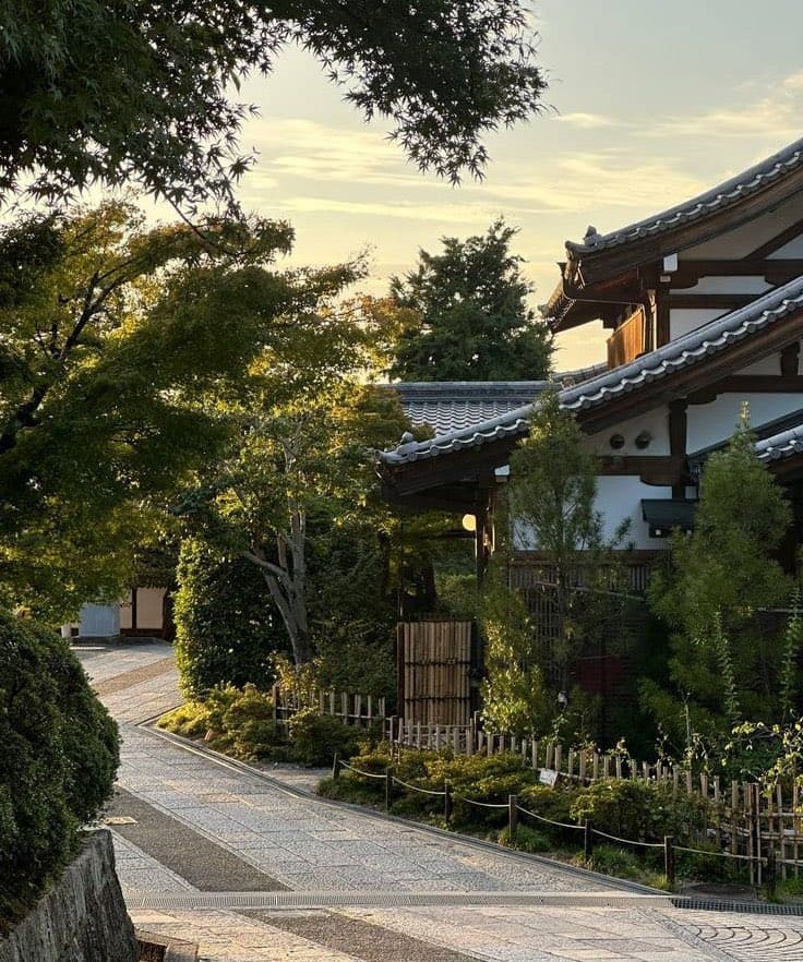 A serene street scene in Kyoto, Japan, featuring a traditional Japanese building with a tiled roof and white walls, surrounded by lush greenery and a stone-paved path.