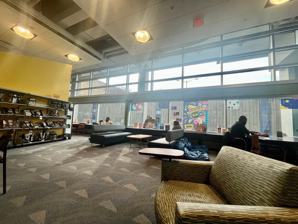 An interior view of a modern public library or community center featuring a seating area with patterned armchairs and a long black sofa. To the left, a yellow wall holds shelves of electronic devices under a sign reading 'GAMES,' while large windows line the back wall with colorful posters displayed on the glass.
