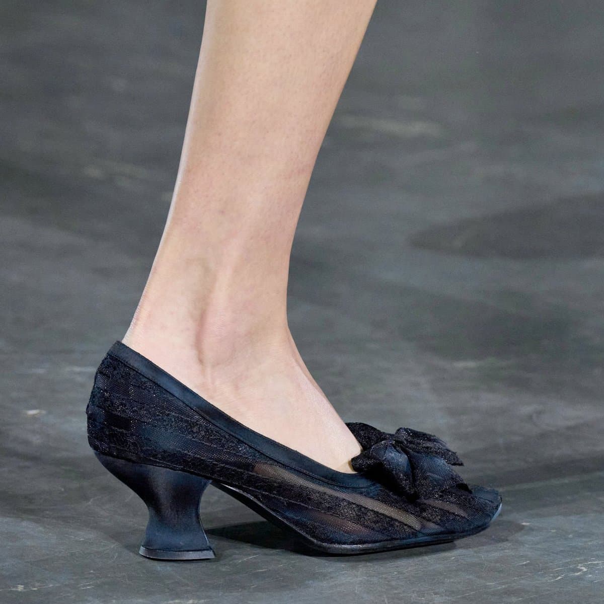 A close-up photograph of a person's foot wearing a black, lace-up heeled shoe with a bow detail, set against a dark, textured floor. The image focuses on the shoe's design and the wearer's lower leg.