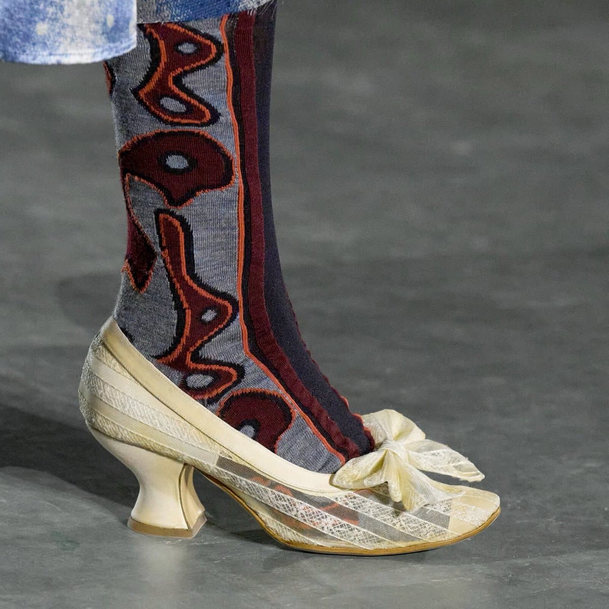 A close-up photograph of a person's lower leg and foot on a runway, wearing a cream-colored, lace-up heeled shoe with a bow and a tall, patterned knit sock. The sock features a bold, abstract design in maroon, orange, and grey.