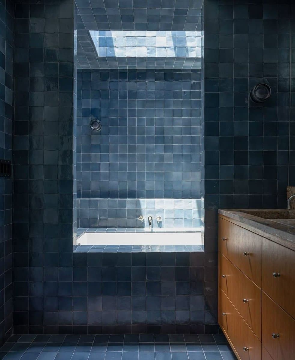 A bathroom interior featuring walls and a floor covered in dark blue square tiles. A recessed alcove contains a white bathtub with chrome fixtures, and a skylight above it lets in bright natural light. To the right, a wooden vanity with a stone countertop is visible.