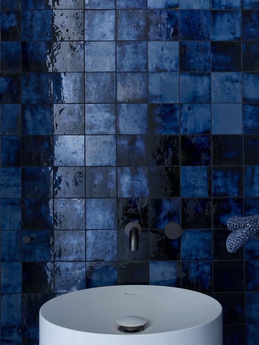 A close-up photograph of a bathroom wall covered in square, blue tiles with a mottled, watercolor-like finish. A white, round vessel sink and a dark metal faucet are visible in the foreground.