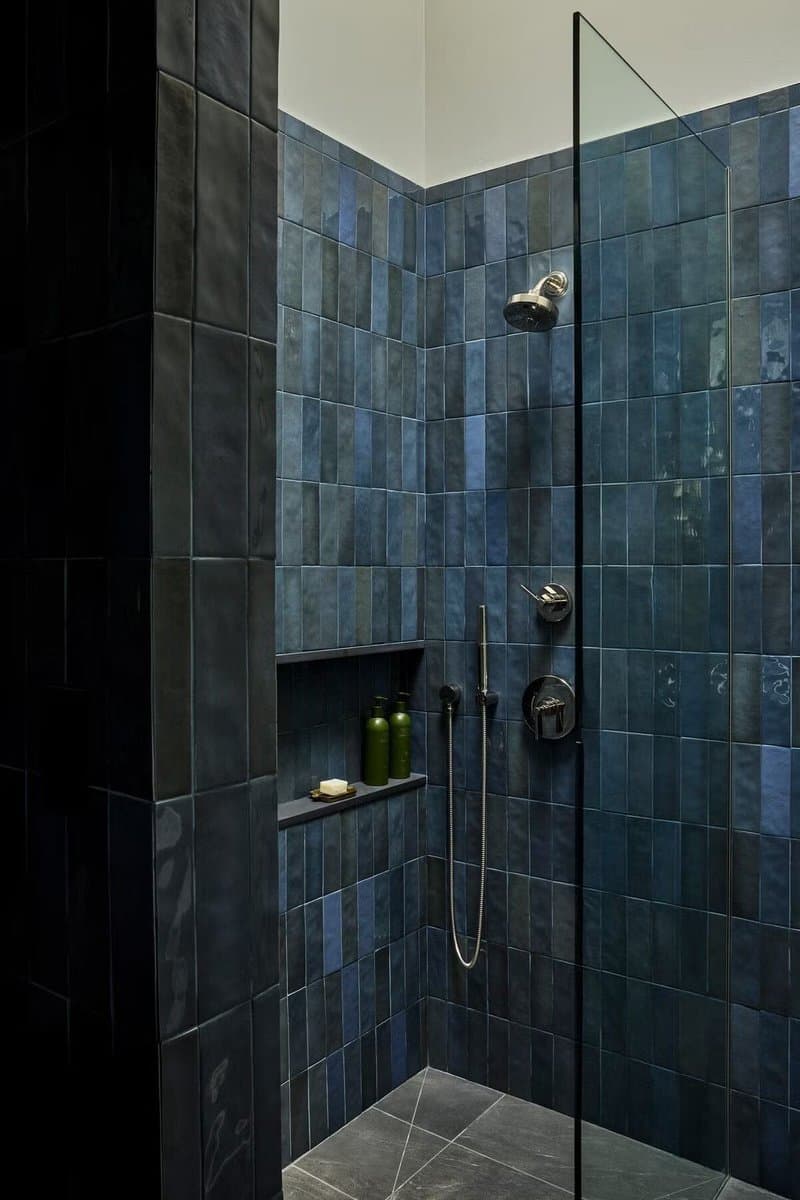 A modern walk-in shower featuring walls covered in glossy, rectangular blue tiles with varying shades. The space includes a glass partition, a chrome shower head and controls, and a built-in niche holding green bottles and a bar of soap.