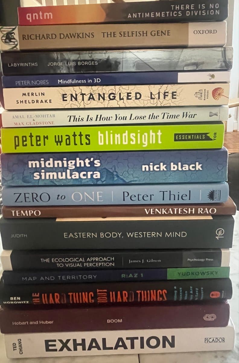 A tall stack of paperback and hardcover books photographed from the side, showing their spines. The collection includes titles on science, philosophy, and fiction, such as 'The Selfish Gene', 'Blindsight', and 'Exhalation'.