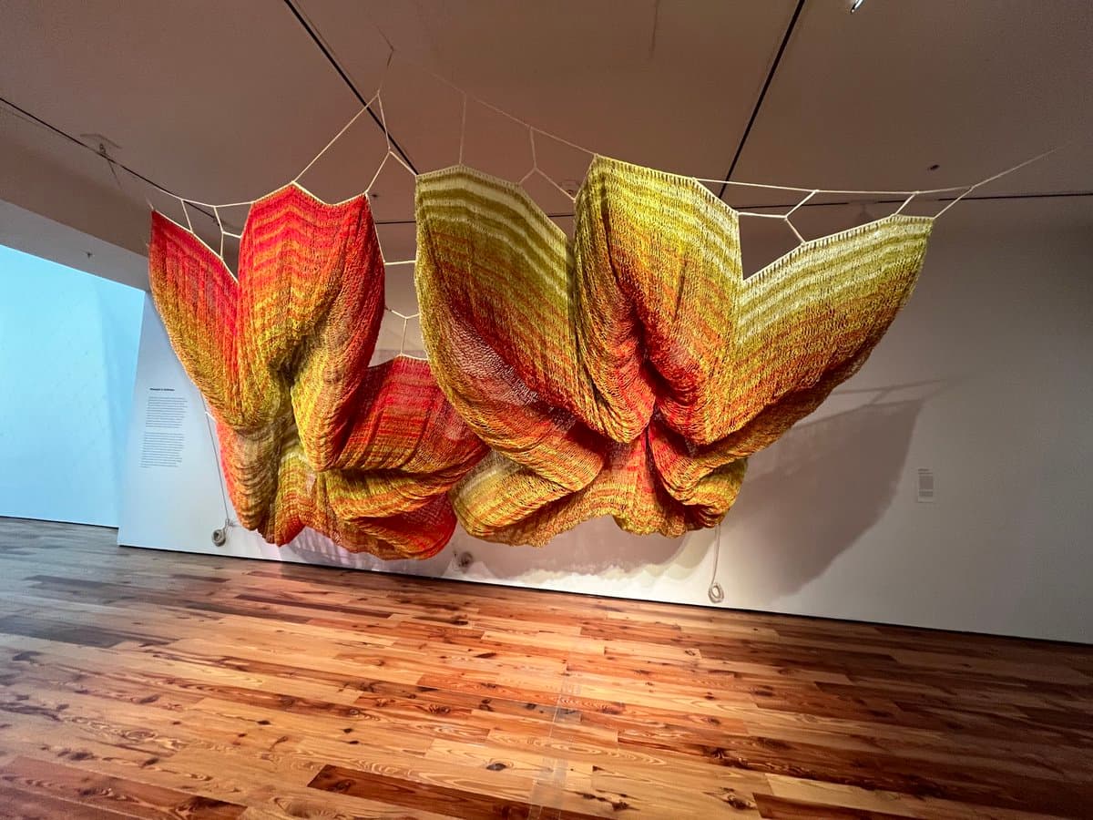 A large, suspended textile art installation featuring a butterfly-like shape in red, orange, and yellow hues hangs from a white net in a gallery with wooden floors.