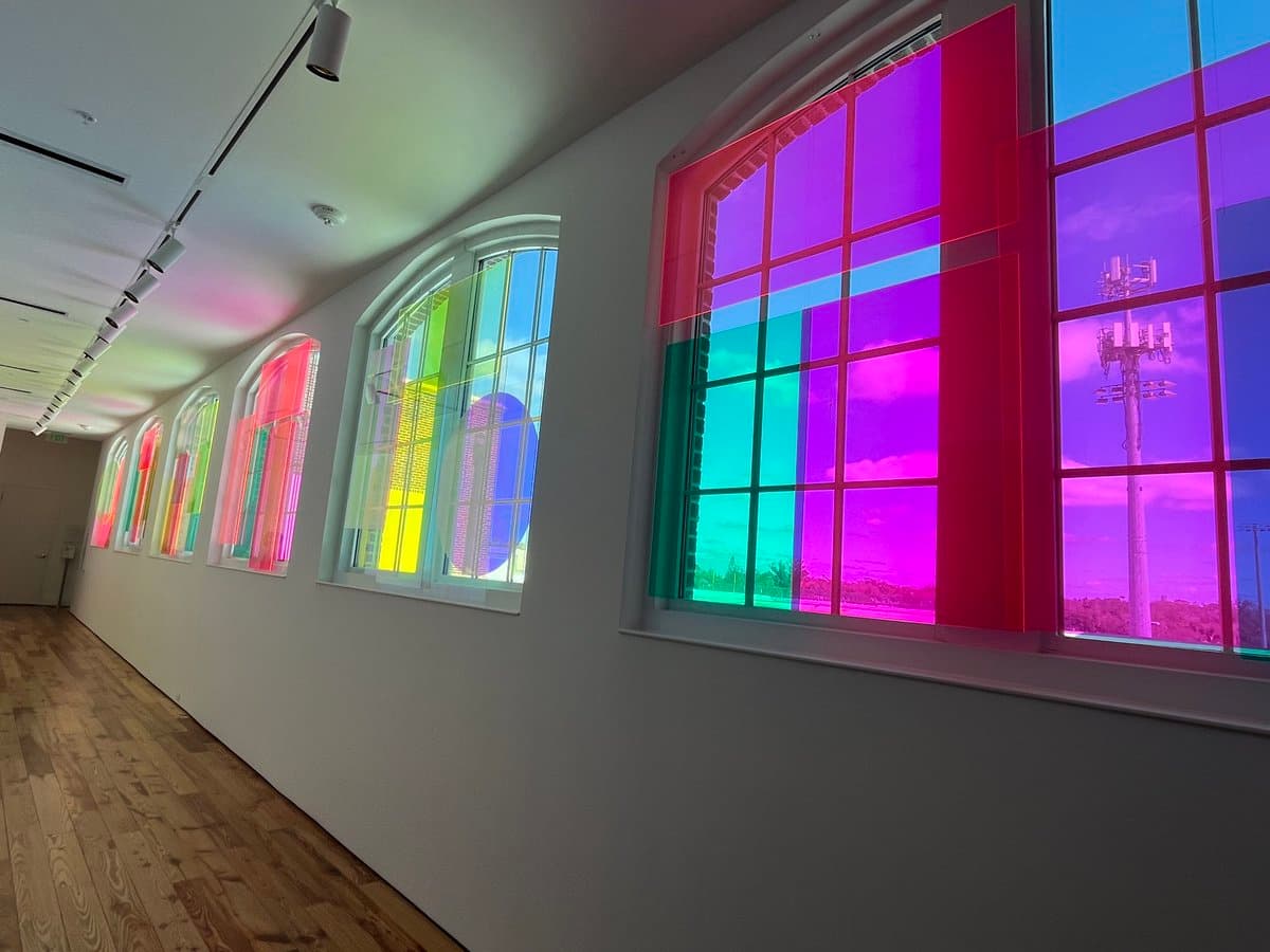 A wide-angle photograph of an art museum gallery featuring a row of large windows covered with vibrant, translucent colored panels in shades of pink, purple, green, and yellow. The interior has white walls and a wooden floor, with track lighting on the ceiling, while the exterior view through the windows shows a bright sky and a telecommunications tower.