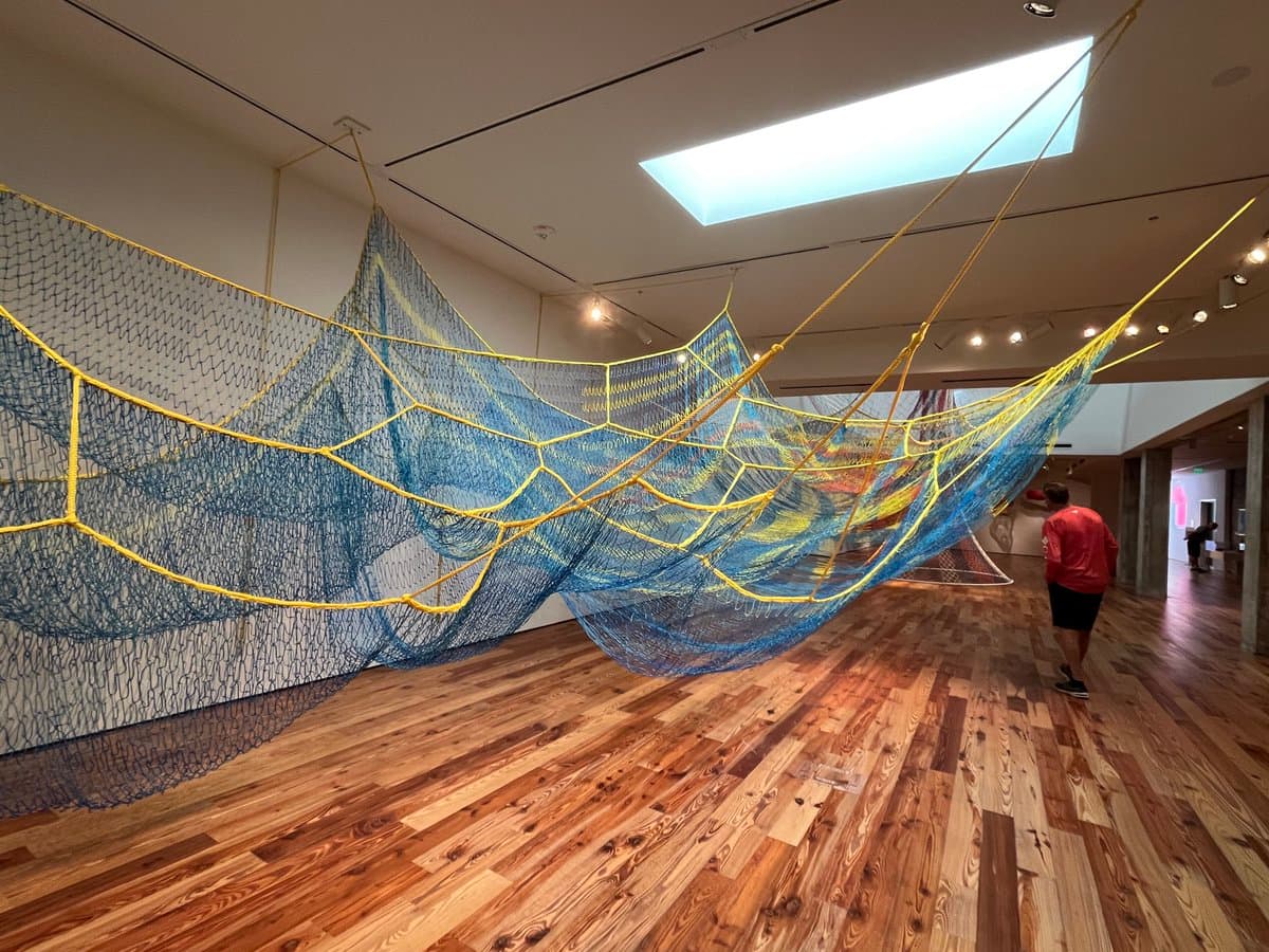 A large, suspended net art installation in blue and yellow hangs from the ceiling of a spacious art museum gallery with wooden floors. A person in a red shirt stands on the right, observing the artwork.
