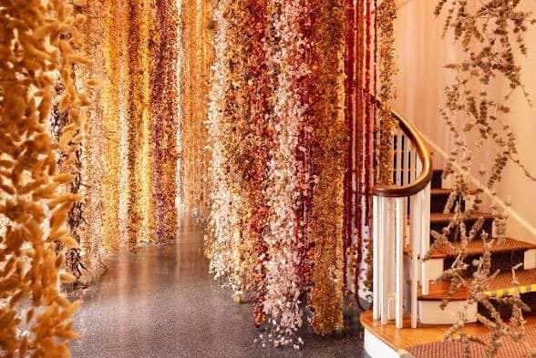 A photograph of an indoor orchid display at the Selby Gardens Orchid Show, featuring numerous vertical strands of dried flowers in warm autumnal colors hanging from the ceiling. A wooden staircase with a white balustrade is visible on the right side of the room.