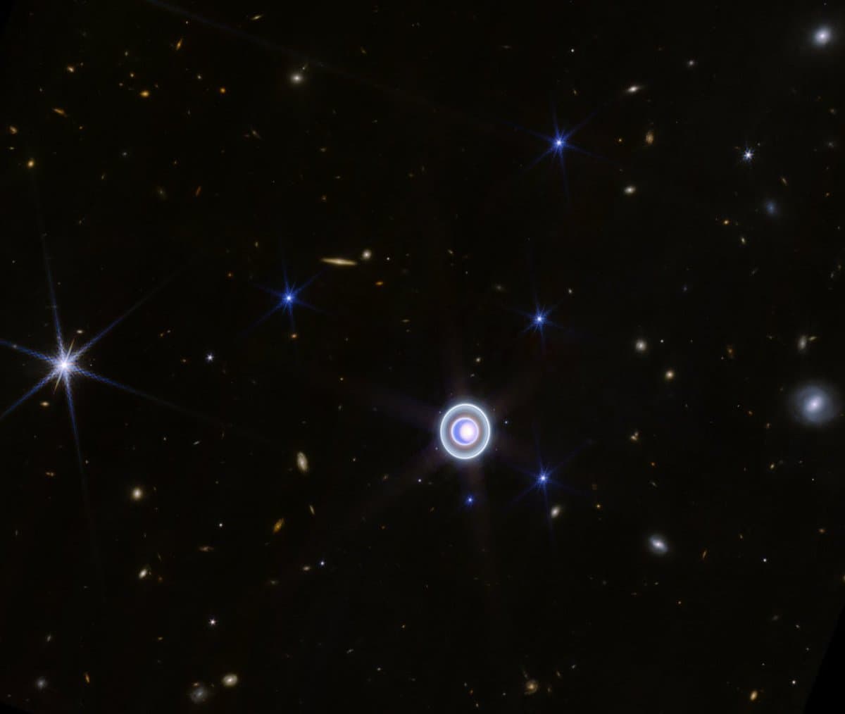 A deep-space photograph showing a field of stars and distant galaxies against a black background. A prominent central object features a bright core surrounded by a distinct, glowing ring, likely a gravitational lensing effect.