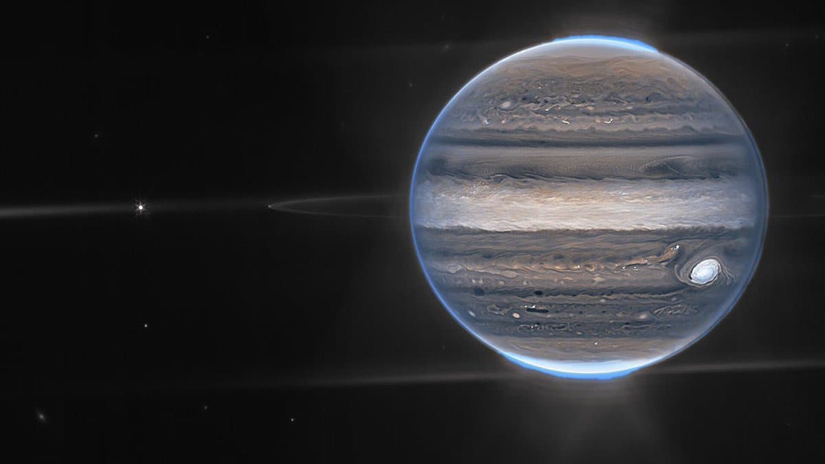 A high-resolution image of the planet Jupiter showing its banded cloud patterns and the Great Red Spot, set against the blackness of space with a bright blue atmospheric glow along its edges. The image features horizontal diffraction spikes extending from the planet, characteristic of the James Webb Space Telescope's optics.