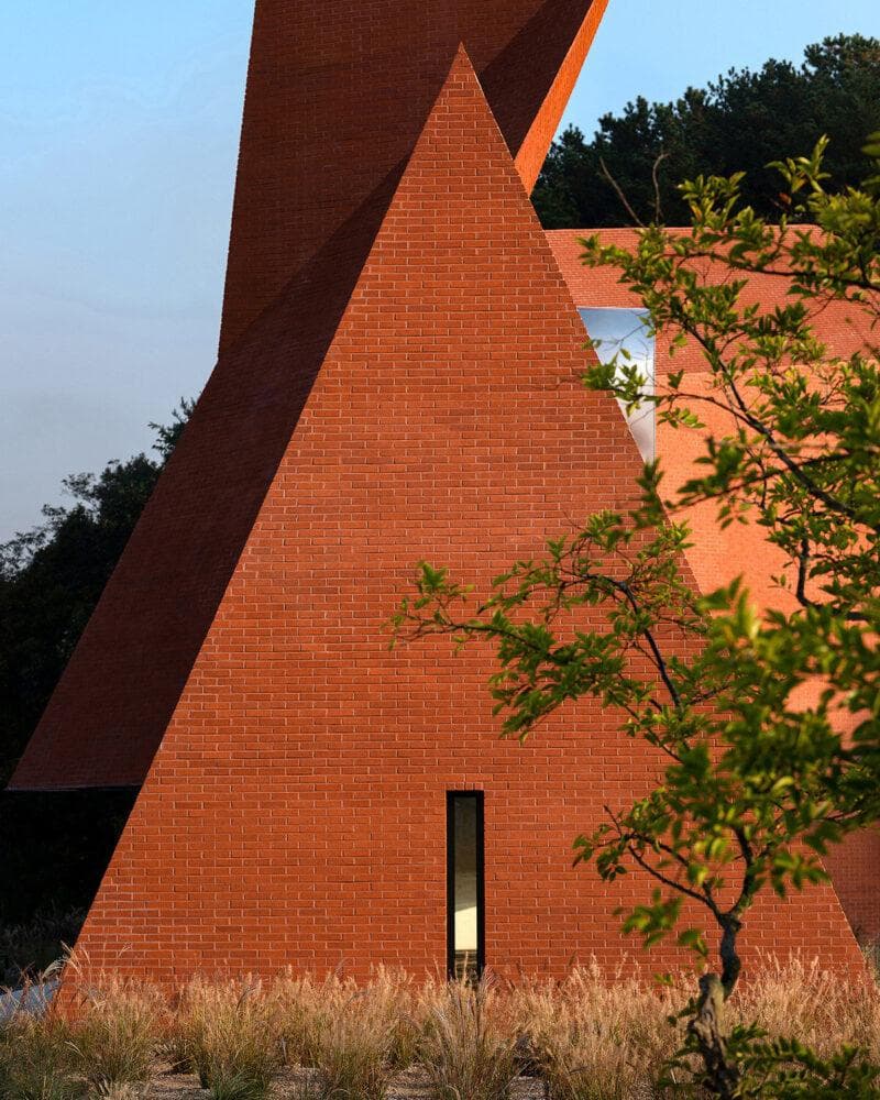 A photograph of a modern architectural structure featuring bold, angular red brick walls and a tall, narrow window, set against a backdrop of trees and a clear sky. A leafy tree branch and tall ornamental grasses frame the foreground.