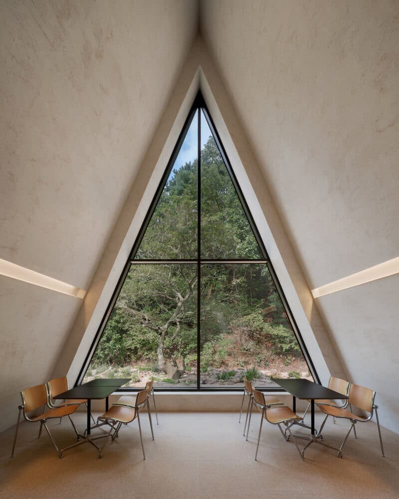 A minimalist interior space featuring a large, triangular window that frames a view of a forest. The room has sloped, textured walls and is furnished with two small tables and chairs arranged symmetrically in front of the window.
