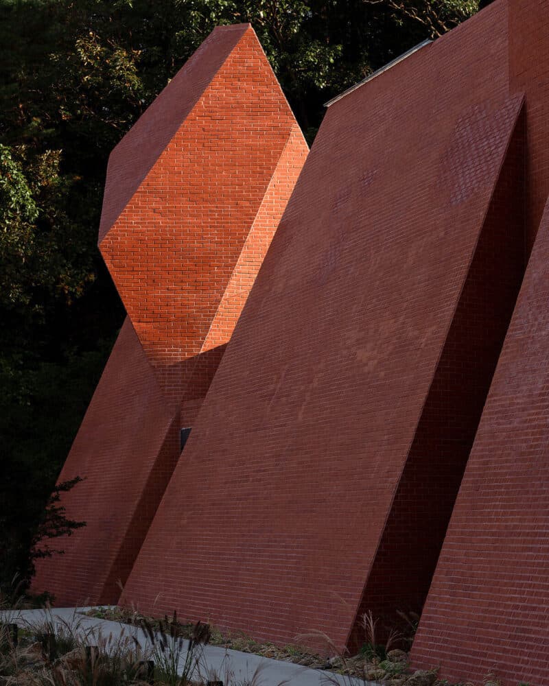 A photograph of a modern architectural structure featuring angular, geometric walls made of red brick. The building is set against a backdrop of dark green trees, with sunlight highlighting the textured brick surfaces and creating deep shadows in the recessed sections.