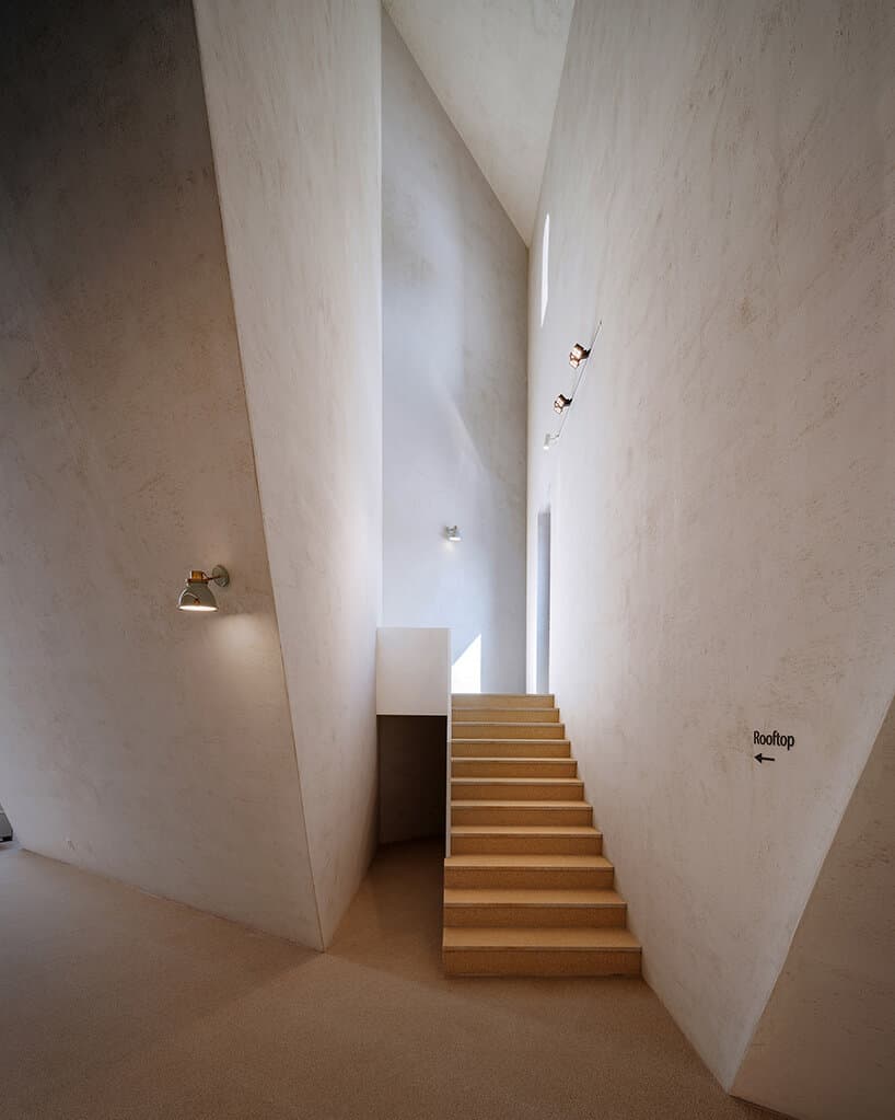A minimalist interior photograph showing a straight staircase with light-colored steps leading up a narrow, white-walled corridor. The space features smooth, textured walls, a small wall-mounted light on the left, and a series of small lights along the right wall. A sign on the right wall indicates the direction to the rooftop.