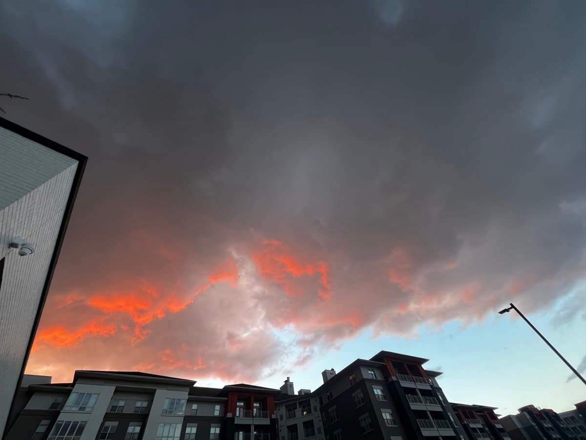 A low-angle photograph of a dramatic sunset sky filled with dark storm clouds illuminated by bright orange and pink light from below. The scene is framed by the tops of modern apartment buildings and a street lamp in the foreground.