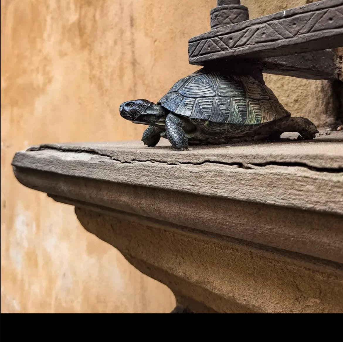 A close-up photograph of a small, dark-colored ceramic turtle figurine with a patterned shell, perched on a carved stone ledge against a textured, ochre-colored wall.