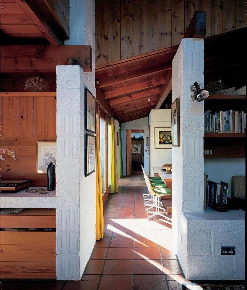 An interior photograph of a house featuring a hallway with terracotta tile flooring, white-painted cinder block pillars, and exposed wooden paneling on the walls and ceiling. The space is furnished with a desk, bookshelves, and green chairs, with sunlight streaming in from a window on the left.