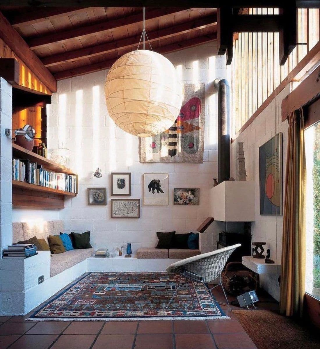 An interior photograph of a rustic living room featuring white brick walls, a wooden beamed ceiling, and a large paper lantern hanging from the center. The room is furnished with built-in bench seating, a patterned area rug, a wire-frame chair, and a fireplace, with various framed artworks and textiles adorning the walls.