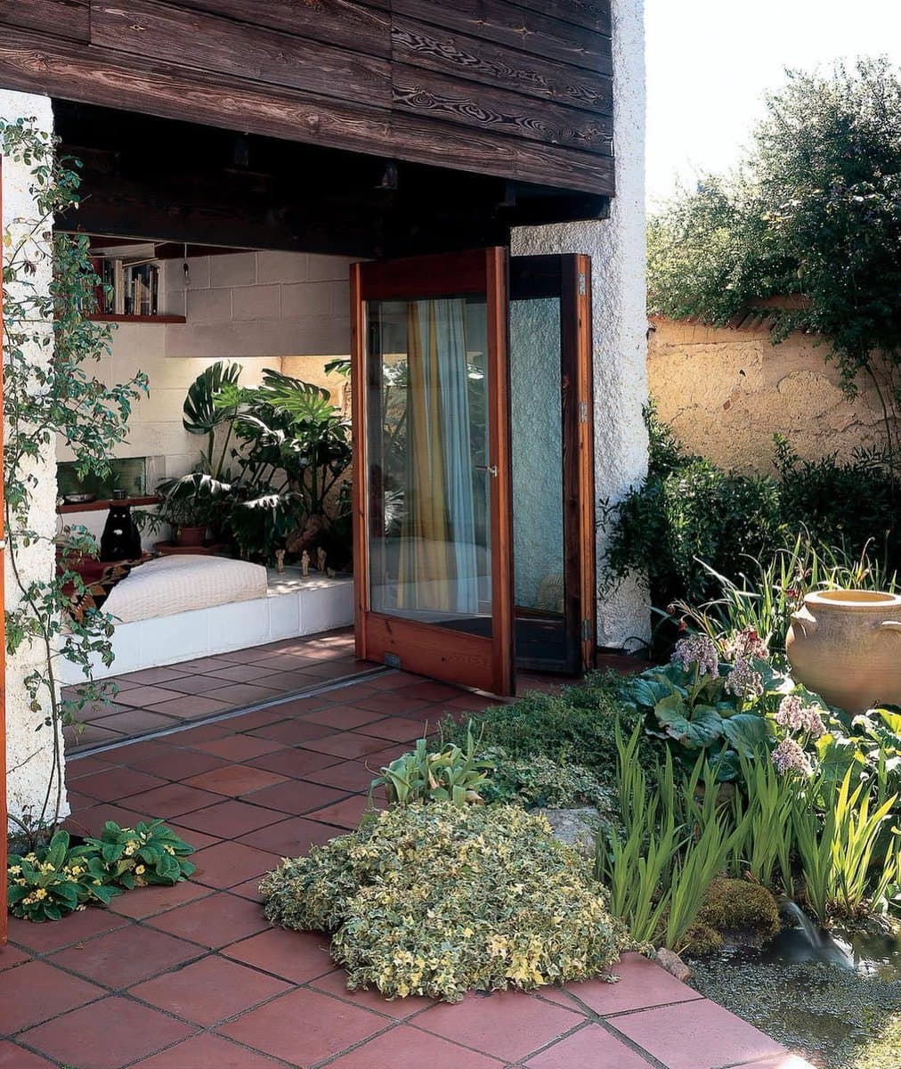 A photograph of a home's exterior featuring a terracotta-tiled patio, a large open wooden-framed glass door, and a lush garden with various plants and a small water feature. Through the open door, a bright interior room with a built-in window seat, houseplants, and a black cat is visible.