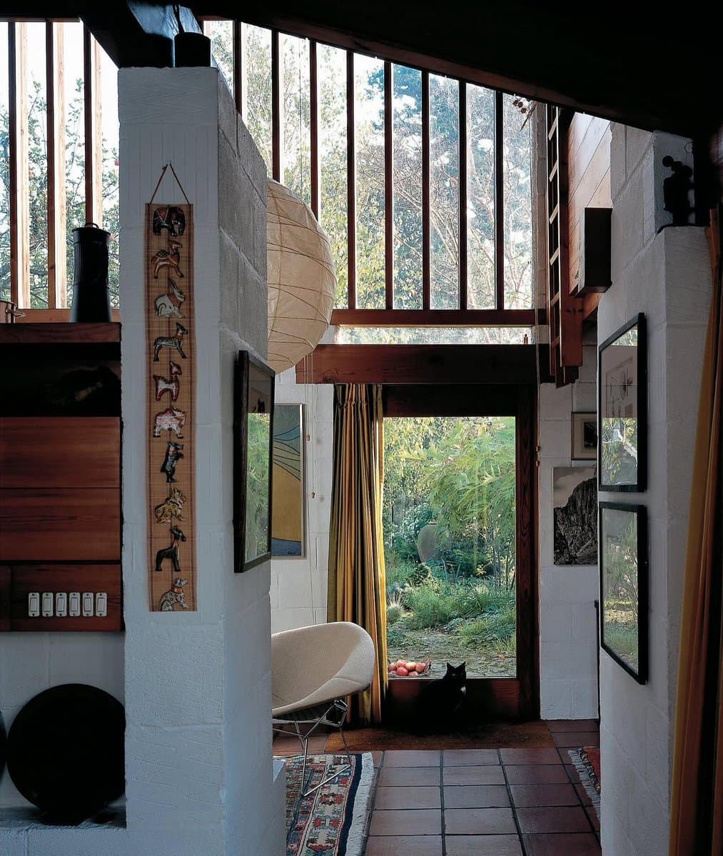 An interior photograph of a modern house featuring white block walls, a large paper lantern, and a tall window looking out onto a garden. A black cat sits on the tiled floor in front of the window, and a vertical wooden art piece depicting animals hangs on the wall to the left.