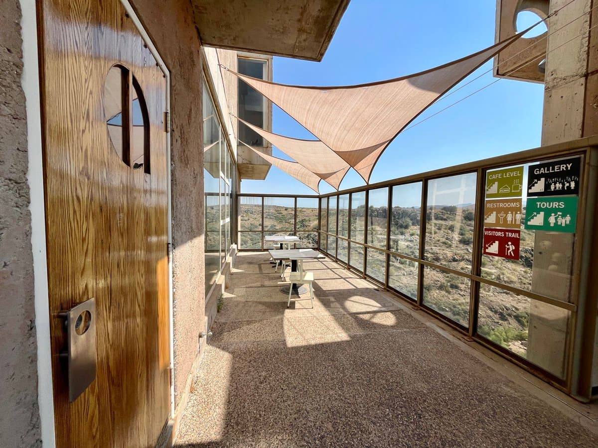 A photo of a concrete and wood balcony with a view of a desert landscape, featuring a large beige shade sail overhead and a set of informational signs on the glass railing.