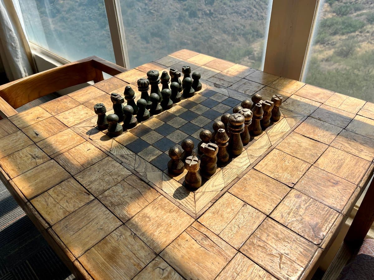 A wooden chess set with carved pieces is arranged on a large, square wooden table that features an inlaid checkerboard pattern. The table sits indoors near a large window, with bright sunlight casting shadows across the surface and a hazy, hilly landscape visible outside.