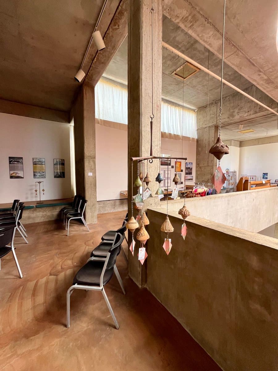 A photograph of a large, open-plan interior space featuring raw concrete pillars and floors. The room contains rows of black chairs, wall-mounted posters, and hanging decorative wind chimes, suggesting a gallery or public building.